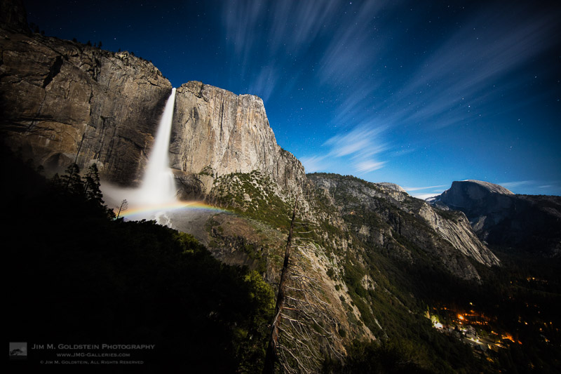 A moonbow spans across upper Yosemite Falls with passing clouds and Half Dome in view.